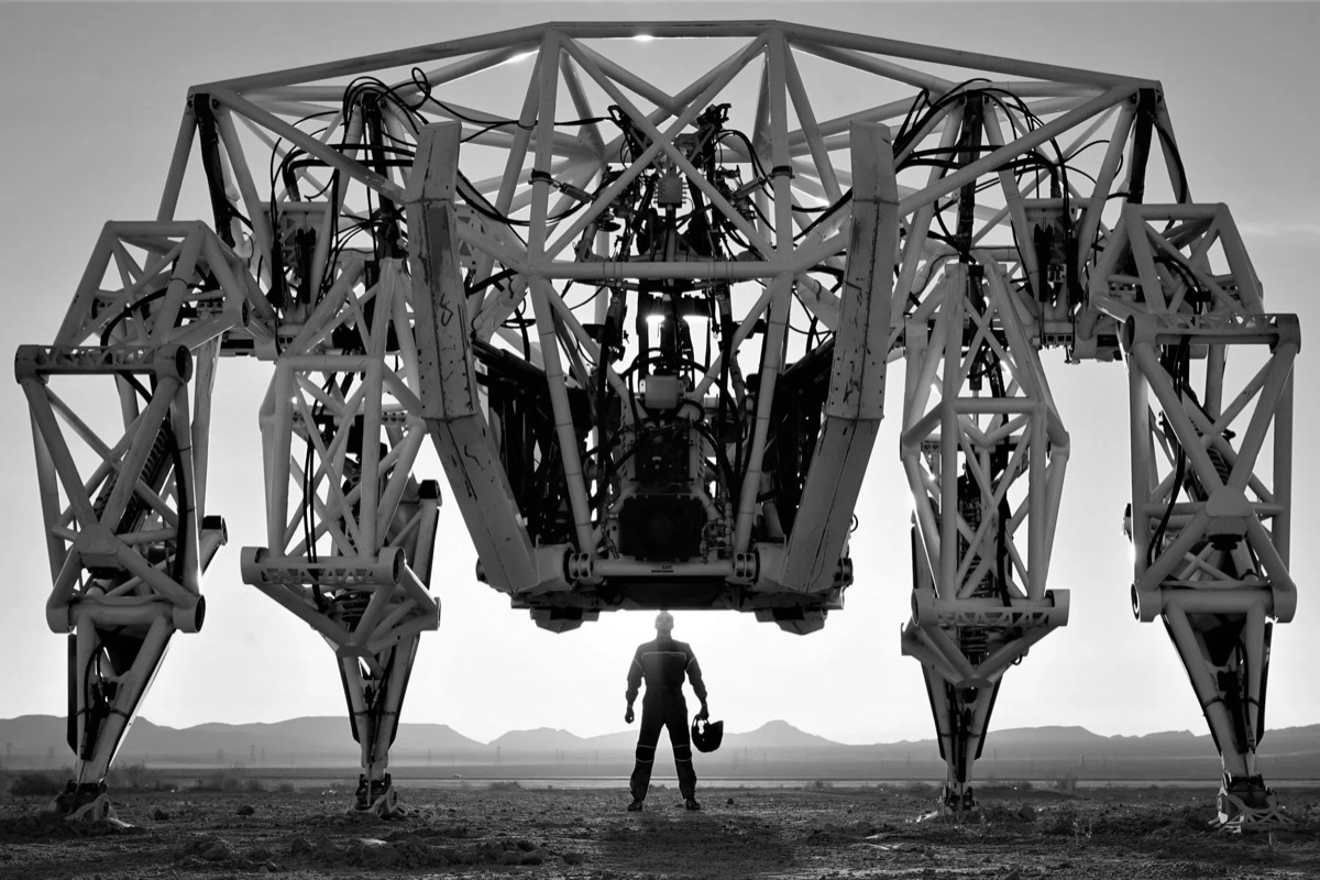 Prosthesis, a giant four-legged walking mech, in black and white at dusk with its pilot standing beneath it